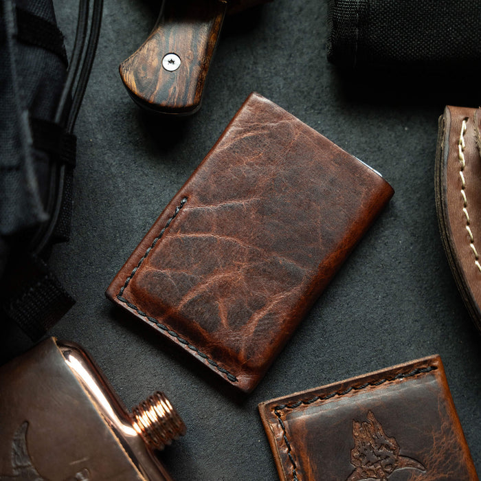MKC Bison Skull mini leather wallet in rich brown leather, surrounded by outdoor gear and tools.