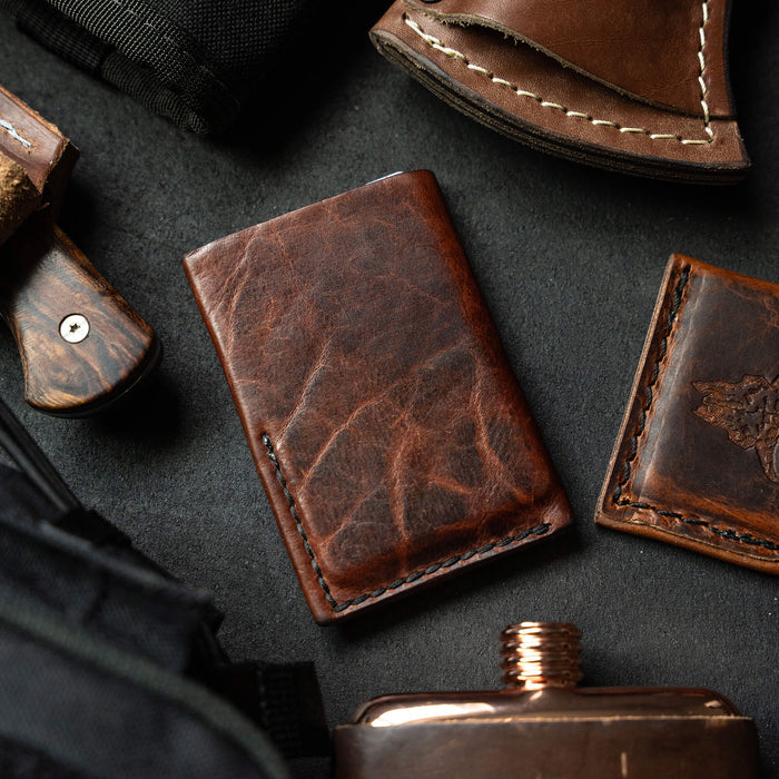 Mini leather wallet featuring a rich brown finish, surrounded by outdoor gear and accessories.