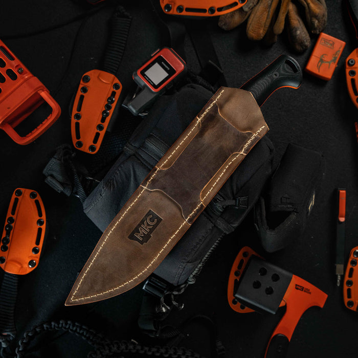 MKC Chopper leather sheath displayed on a tactical bag surrounded by outdoor gear, showcasing its rugged design.
