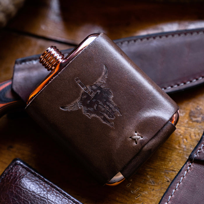 Limited run MKC copper flask with a leather sleeve featuring a bull skull design, resting on a wooden surface.