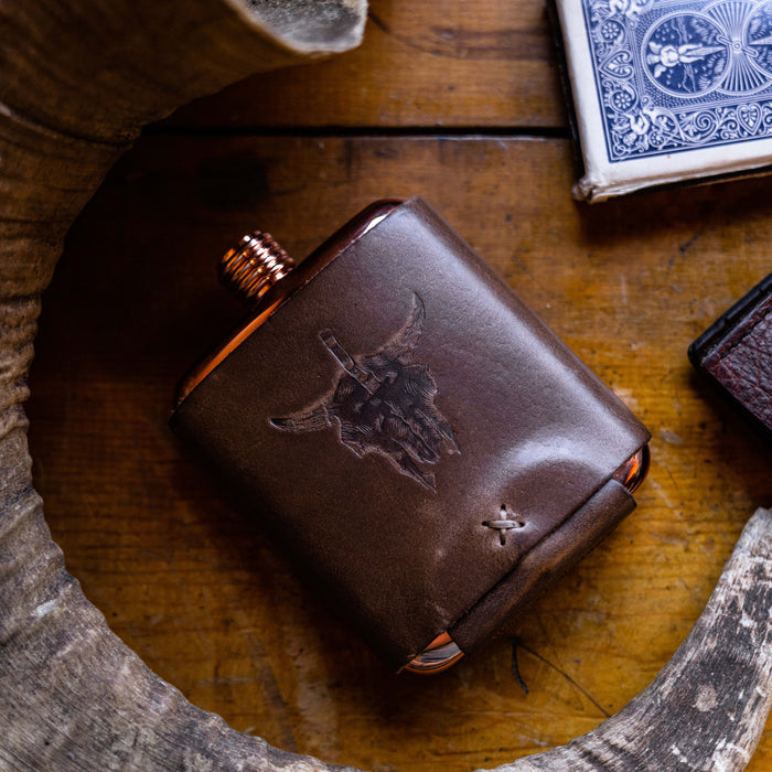 Limited run MKC copper flask with leather wrap featuring a bull skull design, placed on a wooden surface.