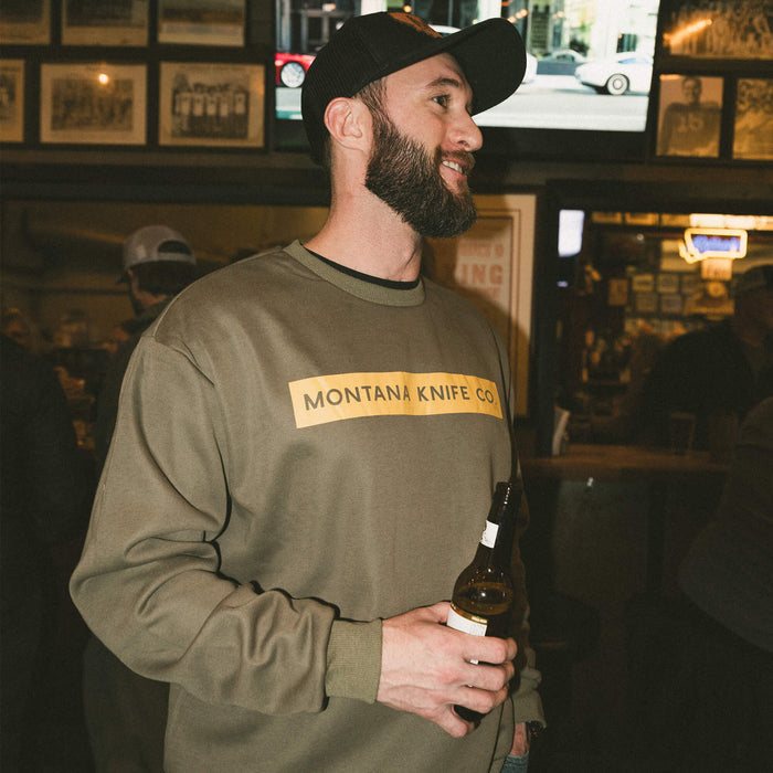 Olive crewneck sweatshirt featuring Montana Knife Company logo, worn by a man holding a beer in a social setting.