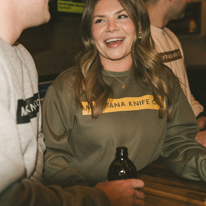Olive MKC crewneck sweatshirt worn by a woman, featuring a bold Montana Knife Company logo, in a casual setting.