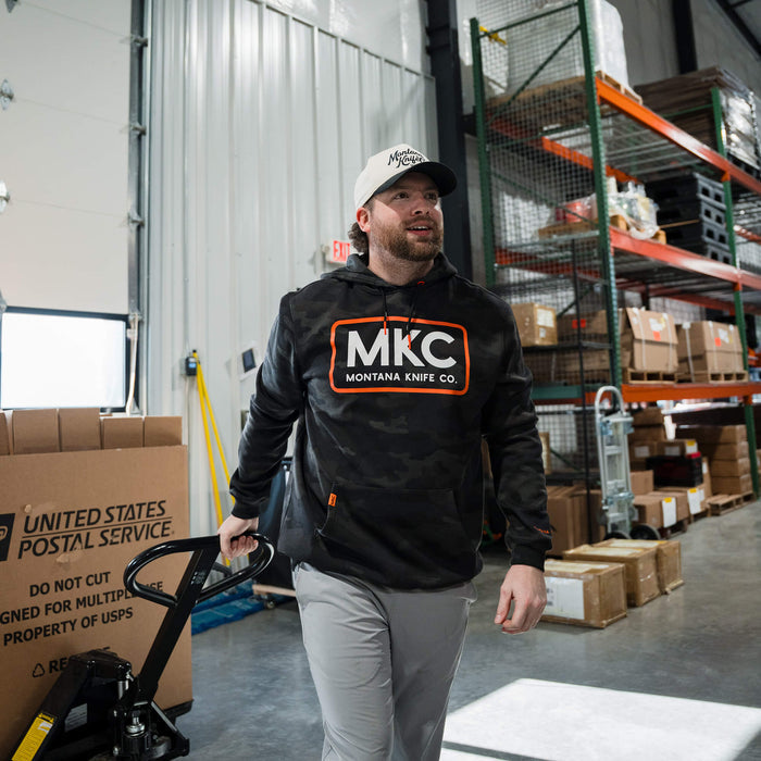 A man wearing a dark woodland camo MKC fleece hoodie walks through a warehouse, carrying a cart.