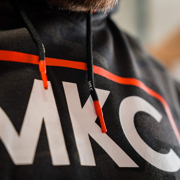Close-up of MKC fleece hoodie in dark woodland camo, featuring a prominent logo and contrasting drawstring details.