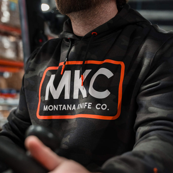 Dark woodland camo fleece hoodie featuring the Montana Knife Company logo in bold orange, ideal for outdoor activities.