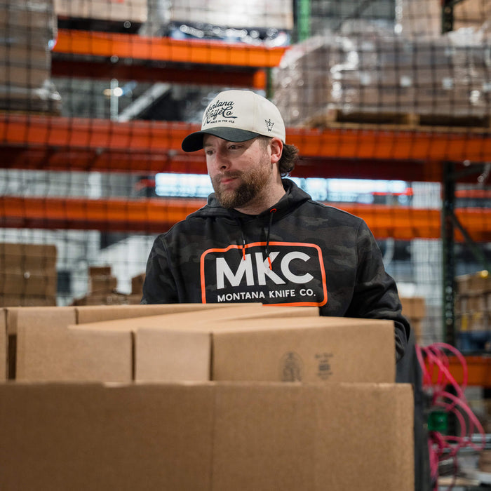 A man wearing a dark woodland camo MKC fleece hoodie stands among stacked boxes in a warehouse.