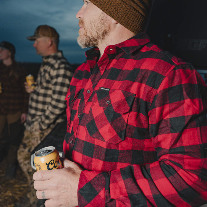 Red and black heavyweight flannel shirt worn by a man holding a can, outdoors with friends in the background.