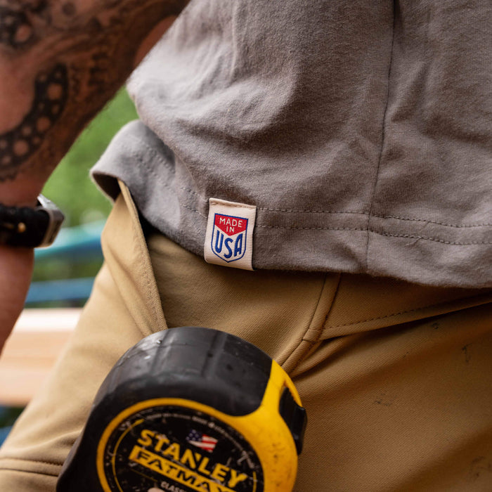 Close-up of a person wearing a stone-colored MKC Heritage Tee with a "Made in USA" tag, holding a measuring tape.
