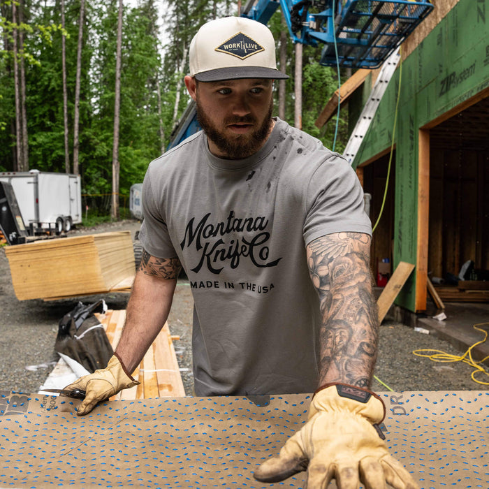 Man wearing a stone-colored MKC Heritage Tee, working outdoors with gloves, showcasing Montana Knife Company branding.