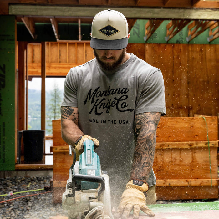 Man wearing a stone-colored MKC Heritage Tee, using a saw in a construction setting, showcasing craftsmanship.