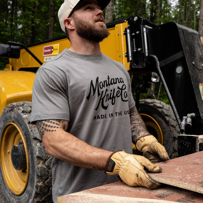 A man wearing a stone-colored Montana Knife Company Heritage Tee stands outdoors, holding a stone while wearing gloves.