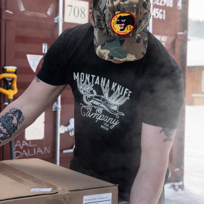 Man wearing a black Montana Knife Company Moose Tee, handling a box outdoors in a snowy setting.