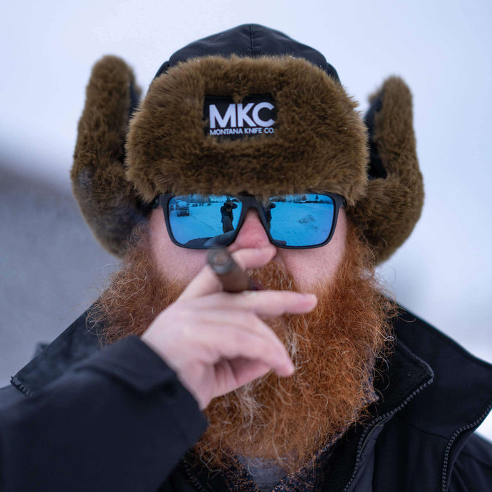 Man wearing an MKC Trapper Hat with fur ear flaps, sunglasses, and a cigar, set against a snowy background.