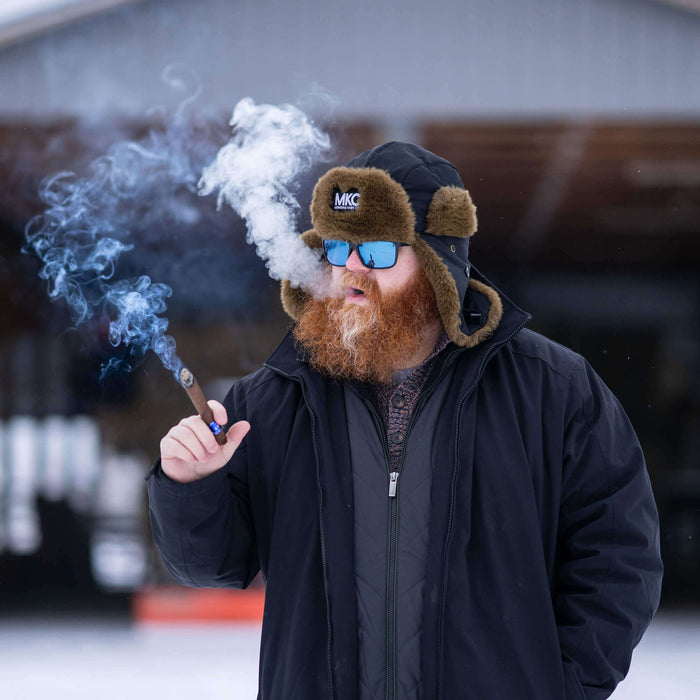 A man with a red beard wearing an MKC Trapper Hat and sunglasses, smoking outdoors in a snowy setting.