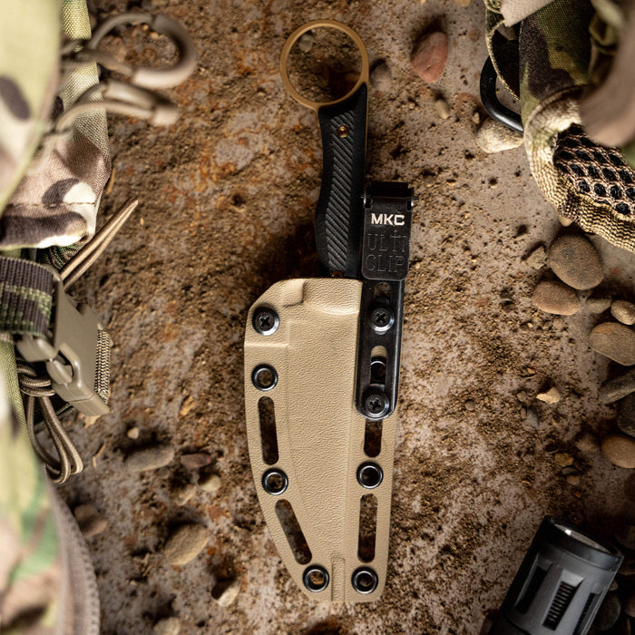 WARGOAT knife with a black handle and tan sheath featuring an MKC clip, set on a textured surface with outdoor gear.