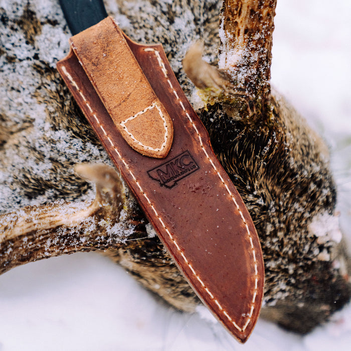 Vertical leather sheath for MKC Whitetail knife, featuring durable stitching and a classic design, resting on a snowy surface.