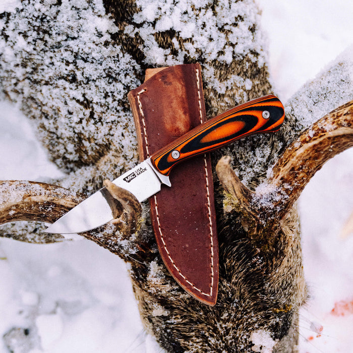 MKC Whitetail knife with orange and black handle in vertical leather sheath on a snowy deer antler backdrop.