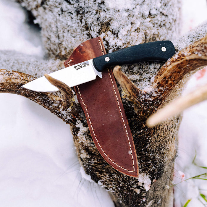 MKC Whitetail knife in a vertical leather sheath resting on a snowy deer antler, showcasing its rugged design.