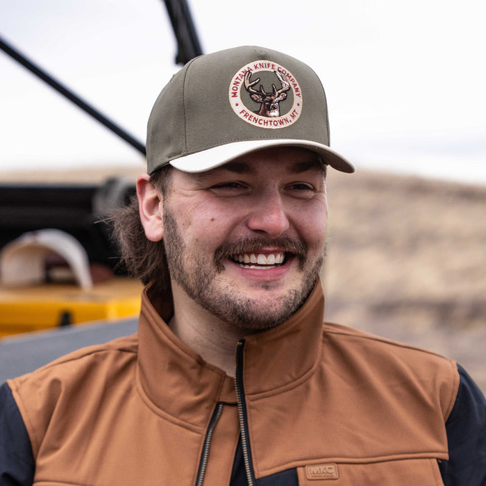 Man wearing a Montana Knife Company Whitetail Hat, smiling outdoors with a rugged landscape in the background.