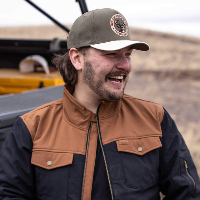 Man wearing a Montana Knife Company Whitetail Hat, smiling outdoors with a rugged jacket and a scenic background.