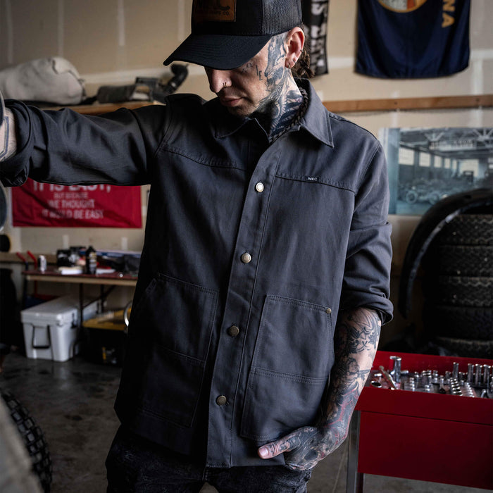 Charcoal MKC Work Jacket made in the USA, featuring a classic design and practical pockets, worn by a tattooed man in a workshop.
