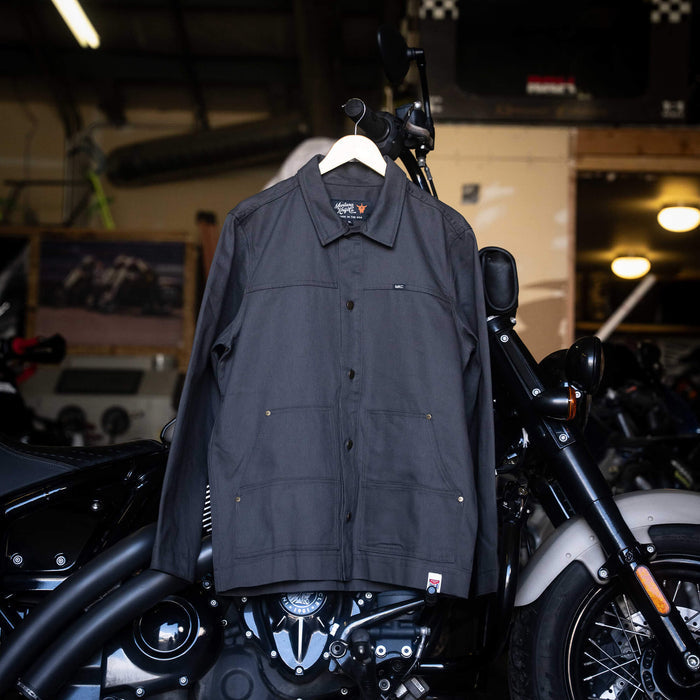 Charcoal MKC Work Jacket displayed on a hanger in a workshop, showcasing its durable design for outdoor professionals.