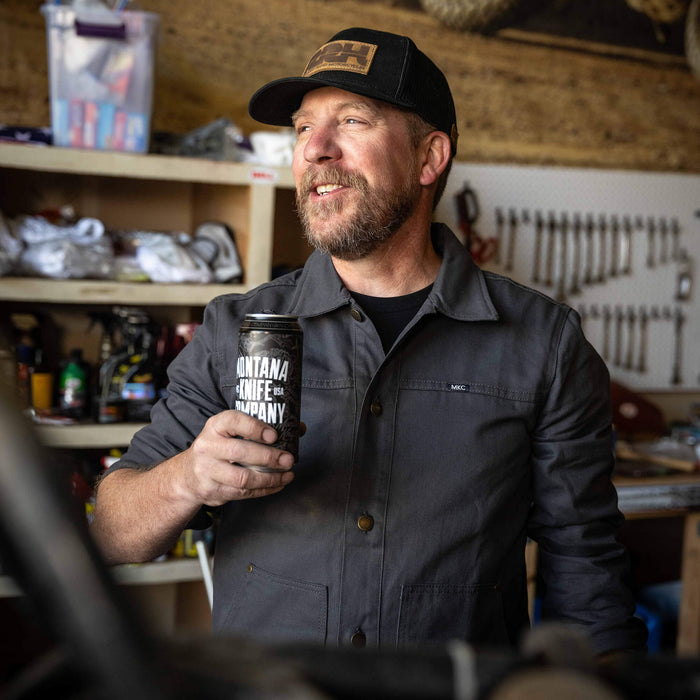 Man wearing a charcoal MKC Work Jacket in a workshop, holding a can with Montana Knife Company branding.
