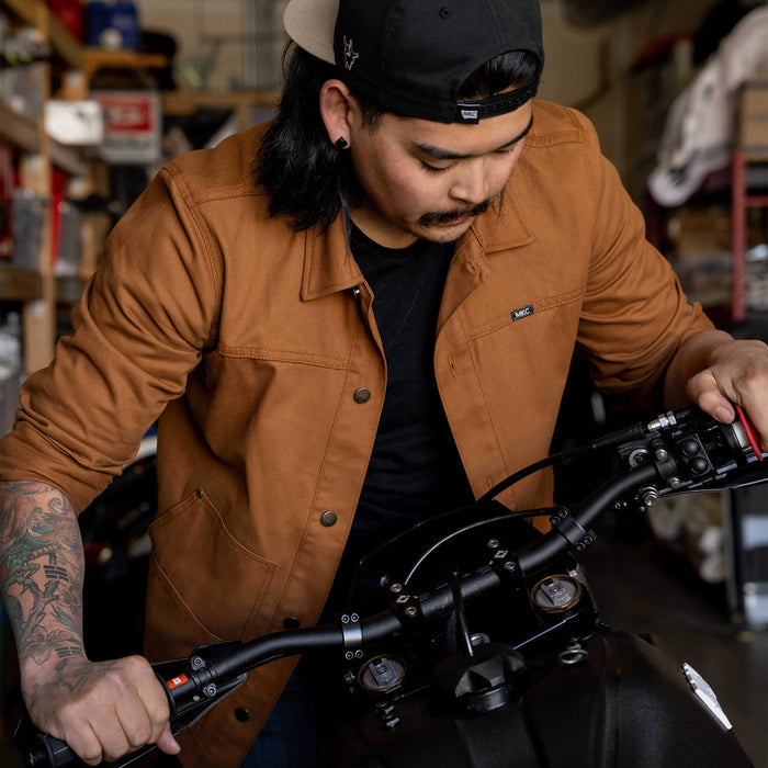 A man wearing a rust-colored MKC work jacket is focused on adjusting the handlebars of a motorcycle in a workshop.