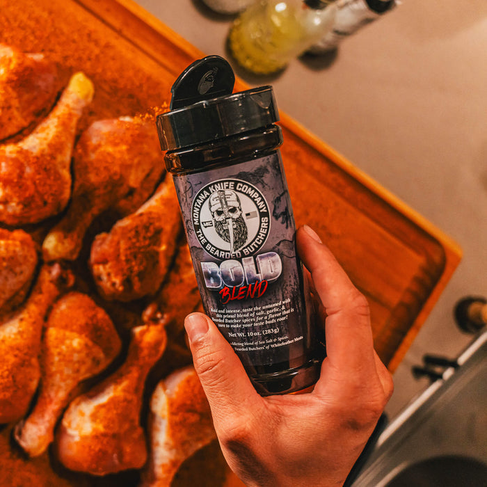 Hand holding a shaker of MKC X Bearded Butchers Bold Seasoning over seasoned chicken legs on a wooden cutting board.