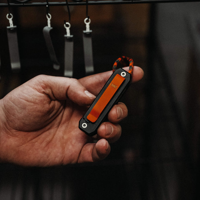 Hand holding the MKC X BIG IDEA DESIGN Ti EDS II, a compact, durable EDC tool with an orange accent and paracord.
