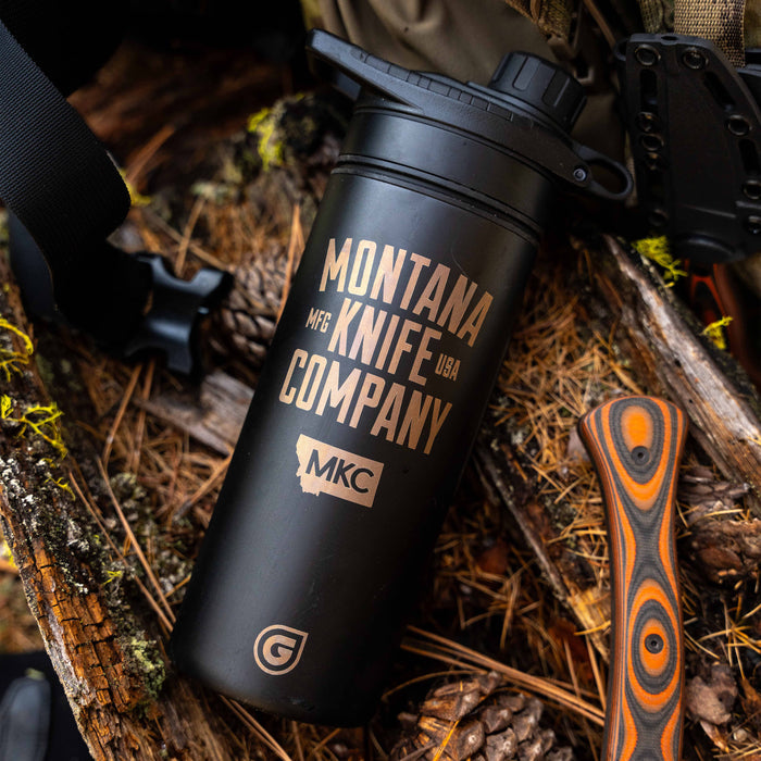 MKC X GRAYL 24oz Geopress Ti Purifier in black with gold branding, placed on a forest floor with a knife handle nearby.