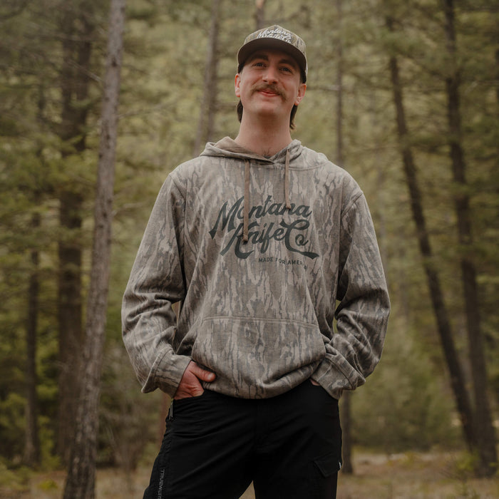 Model wearing a Montana Knife Company x Mossy Oak washed hoodie in Bottomland camo, set in a forested outdoor environment.