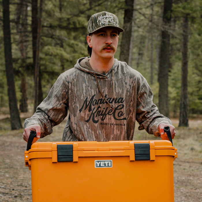 Man wearing a camo hoodie carrying a YETI cooler in a forest setting, showcasing outdoor gear and rugged style.