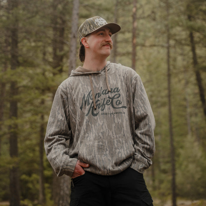 Man wearing a camo hoodie with Montana Knife Company logo in a forest setting, showcasing outdoor style and comfort.