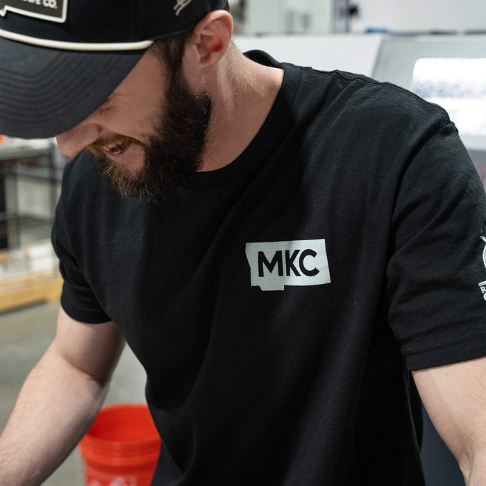 Black MKC x Rocky Mt. Goat Alliance tee featuring a silver MKC logo, worn by a man in a workshop setting.