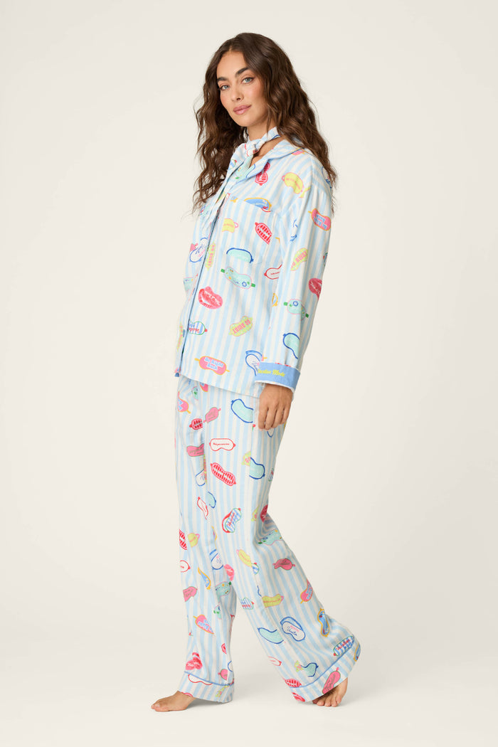 A woman with long wavy hair models the P.J. Salvage Out of Office Eyemask Print 3-Piece Flannel PJ Set—loose, light blue pajamas adorned with colorful illustrations of lips, toothbrushes, and toothpaste. She stands barefoot against a plain light background, looking toward the camera.
