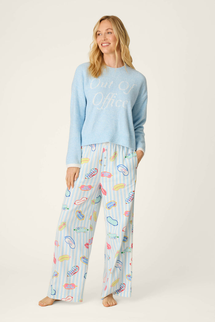 A woman smiles while wearing the P.J. Salvage Out of Office Snuggle Sweater in light blue and pajama pants with a colorful macaron print, standing barefoot against a plain light background.