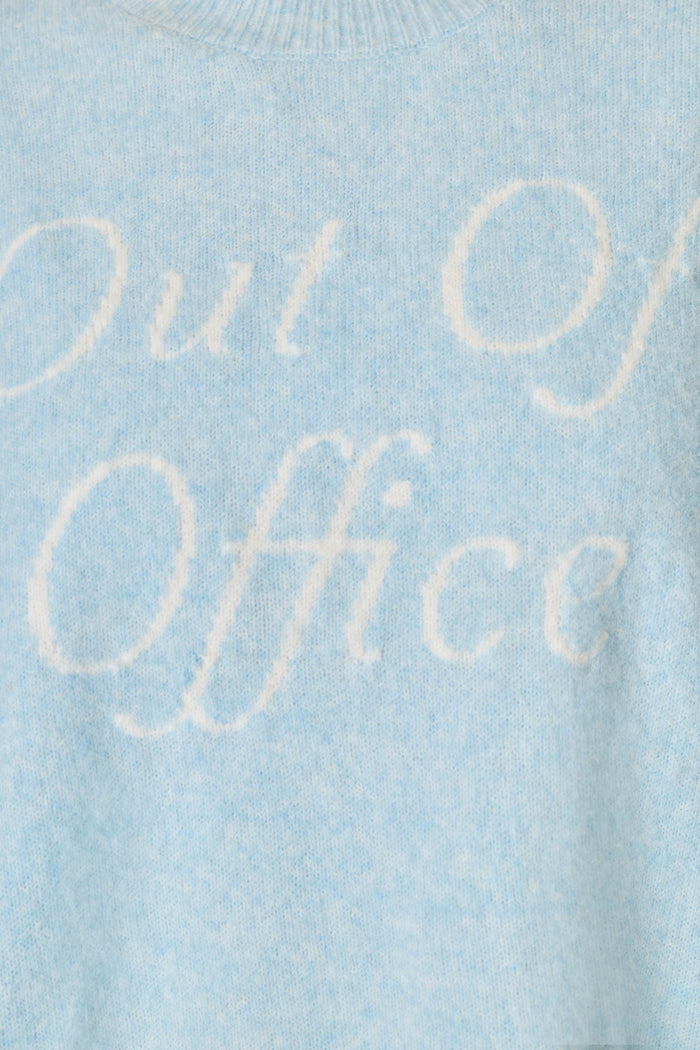 Close-up of a light blue knit sweater with the words 'Out of Office' knitted in a contrasting white script across the chest.