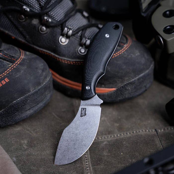 Black PACKOUT SKINNER knife with a curved blade, resting on a rugged surface near outdoor footwear and gear.