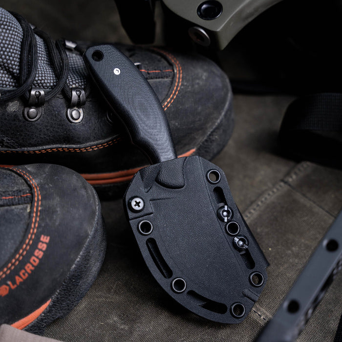 Black PACKOUT Skinner knife resting beside rugged boots, showcasing its durable design for outdoor use.