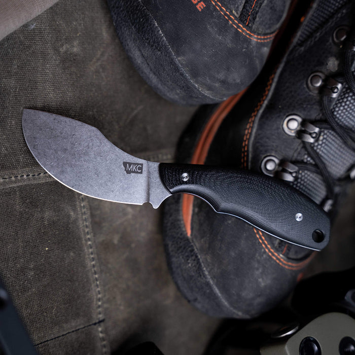 Montana Knife Company's PACKOUT SKINNER in black, featuring a curved blade and ergonomic handle, placed beside rugged footwear.