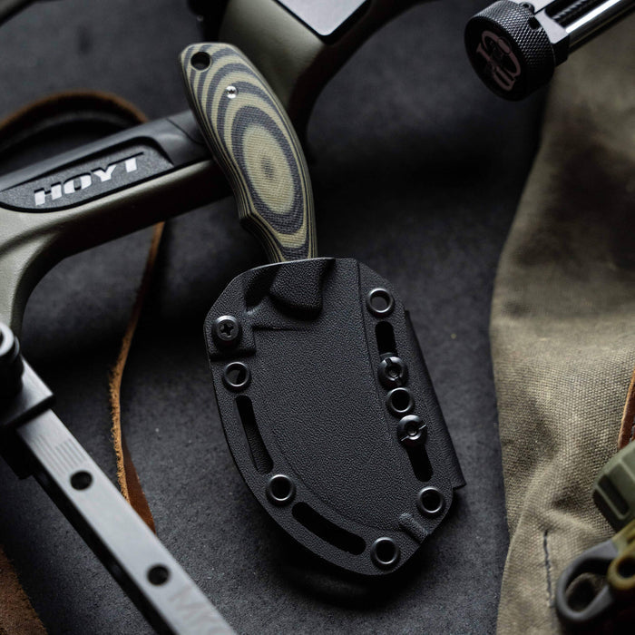 Green and black PACKOUT SKINNER knife with a textured handle, positioned on a dark surface alongside outdoor gear.