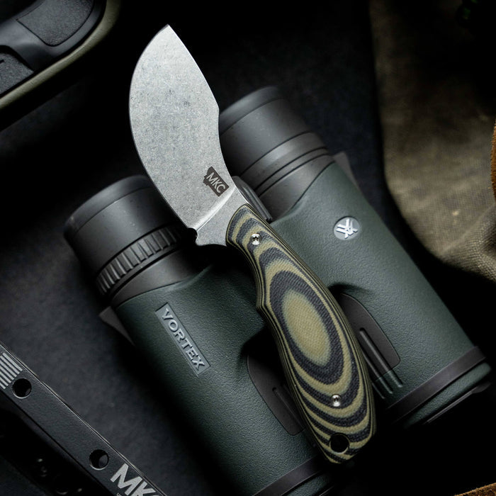 Green and black PACKOUT SKINNER knife rests on binoculars, showcasing its durable blade and ergonomic handle design.