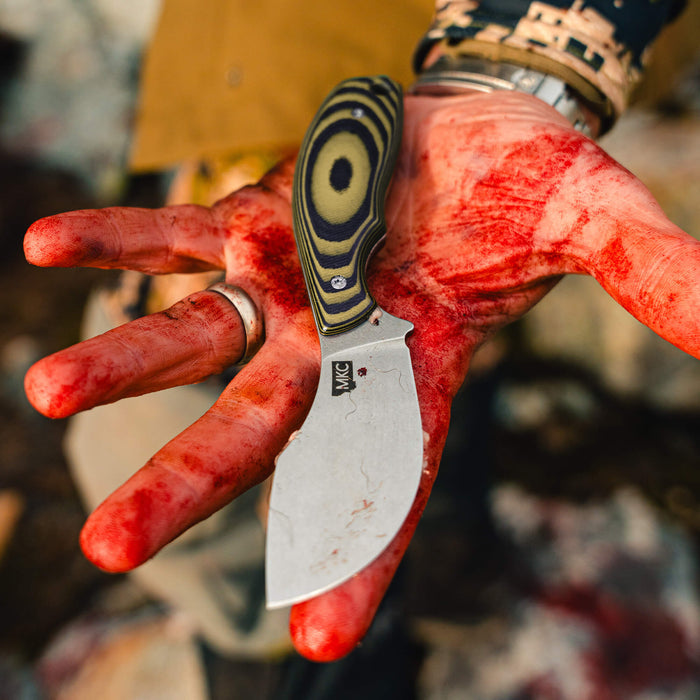 Green and black PACKOUT SKINNER knife held in a blood-stained hand, showcasing its curved blade and ergonomic handle.