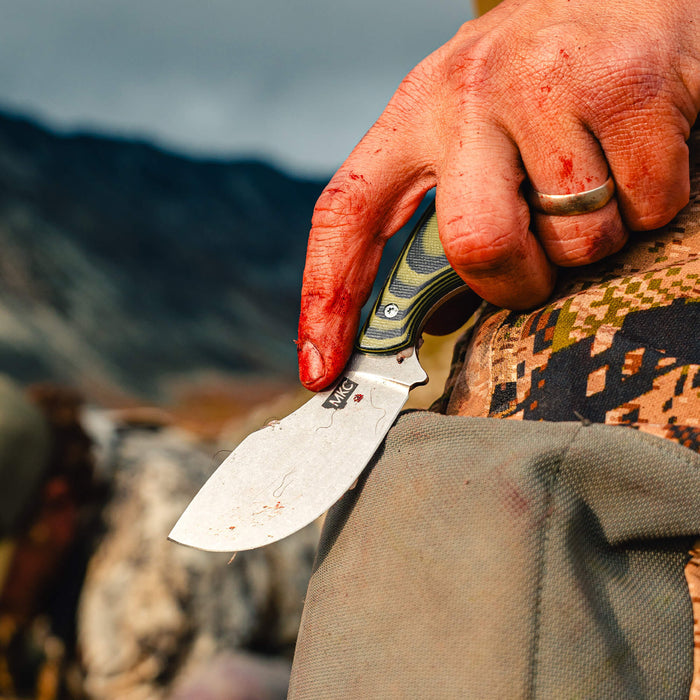 A hand with blood-stained fingers holds the PACKOUT SKINNER knife featuring a green and black handle, set against a rugged outdoor backdrop.