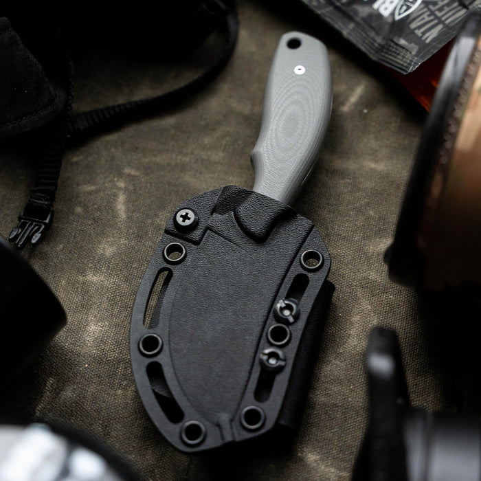 Grey PACKOUT SKINNER knife in a black sheath, surrounded by outdoor gear, showcasing its rugged design for hunting.