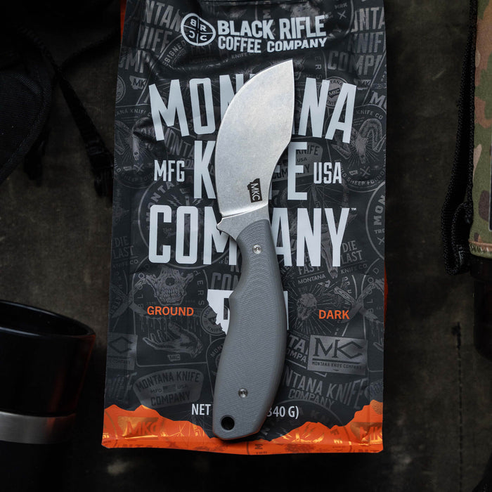 Montana Knife Company's PACKOUT SKINNER in grey, featuring a curved blade and ergonomic handle, displayed on a coffee bag.
