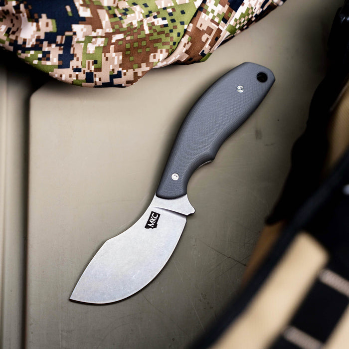 Grey PACKOUT SKINNER knife with a curved blade, resting on a textured surface among camouflage gear.