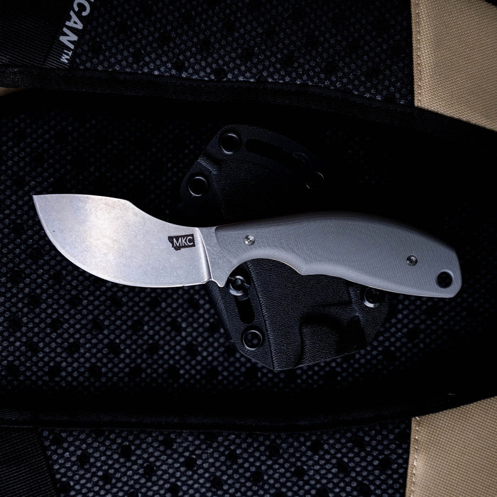 Montana Knife Company's PACKOUT SKINNER in grey, featuring a curved blade and ergonomic handle, displayed on a textured background.